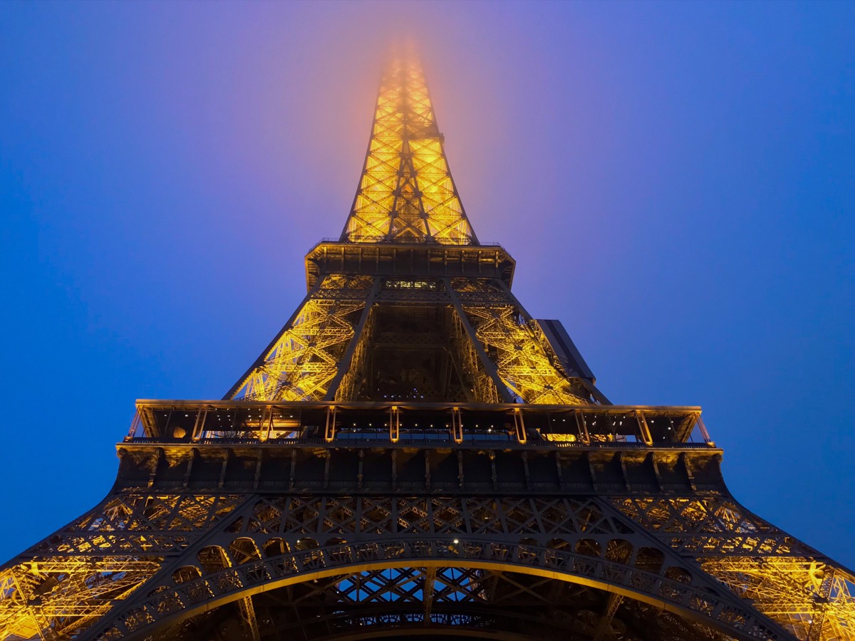 The illuminated Eiffel Tower at blue hour, its top dissolving into fog, seen from below